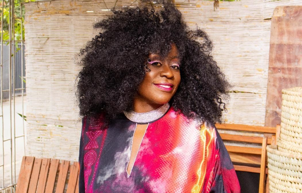 Person with voluminous curly hair wearing a colorful patterned top, smiling outdoors by wooden chairs