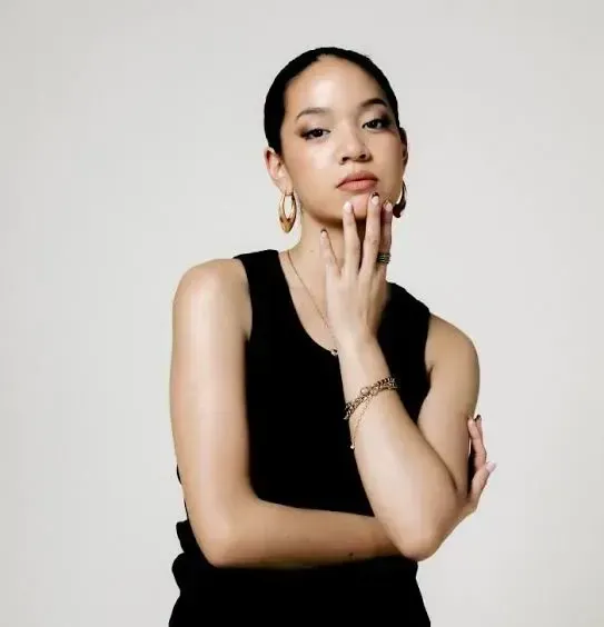 Person in a black sleeveless top posing against a light background, hand resting on chin.