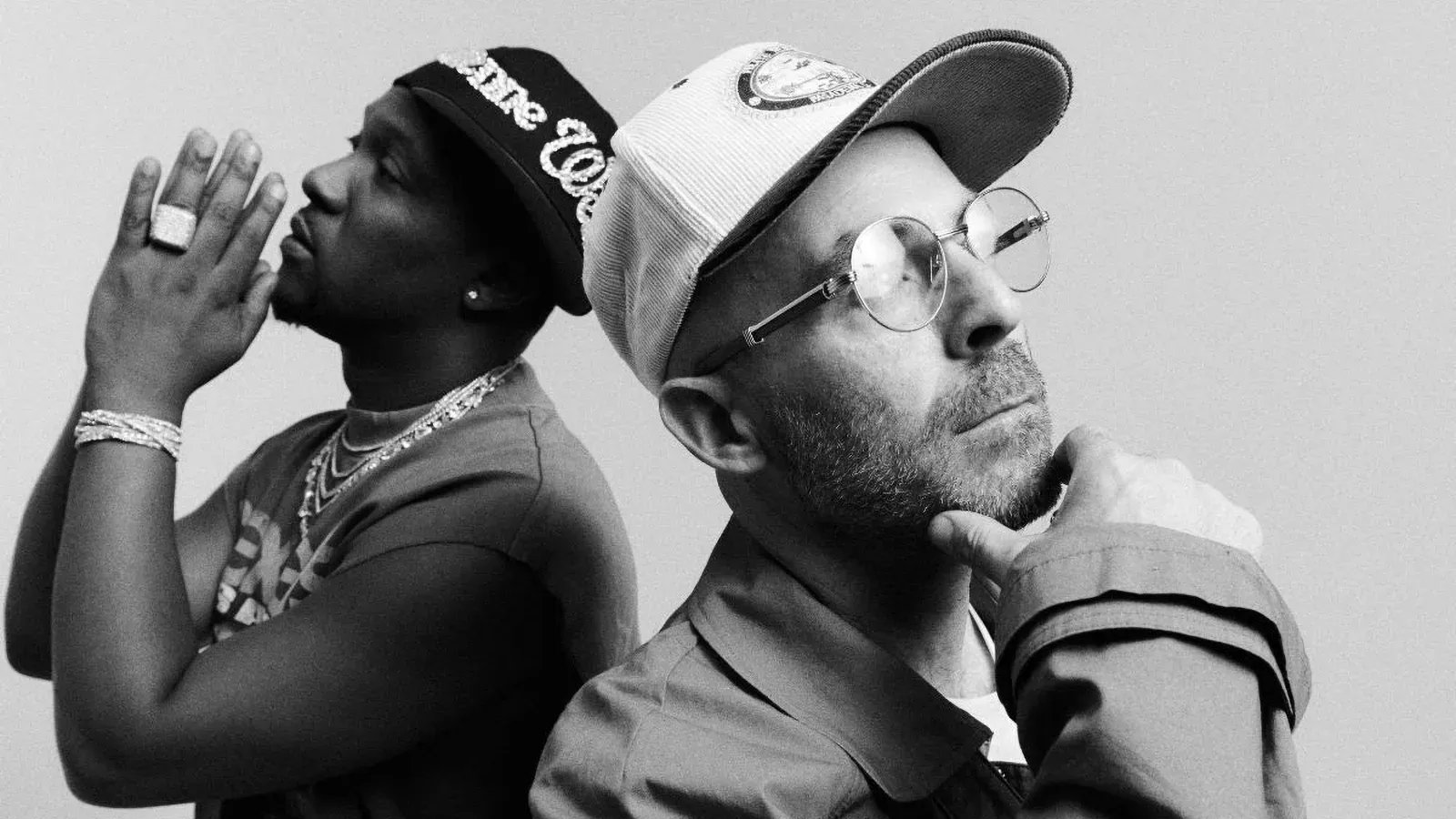 Two men posing back-to-back in black-and-white, one in sunglasses and a cap, one in a tank top and beanie.