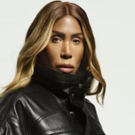 Portrait of a person in a black leather jacket and high collar, with long blonde hair on a light background