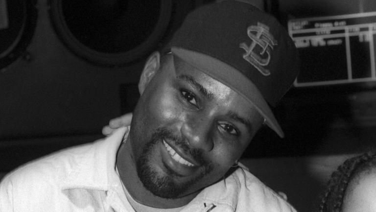 Black-and-white portrait of a smiling man in a cap, leaning toward the camera in a dimly lit setting