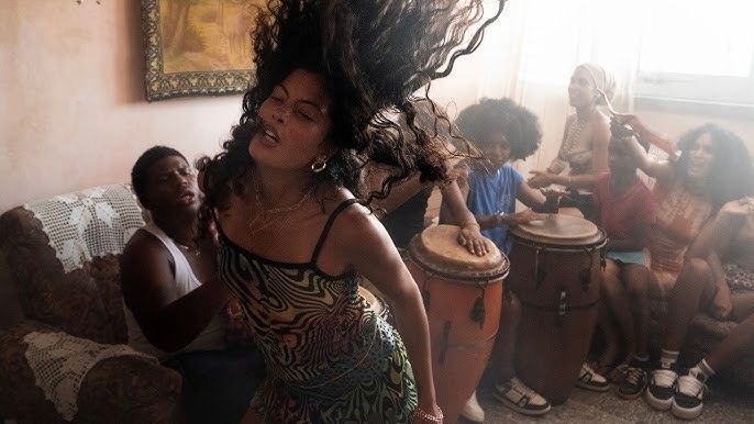 Woman dancing in a living room as people sit and play drums in the background.