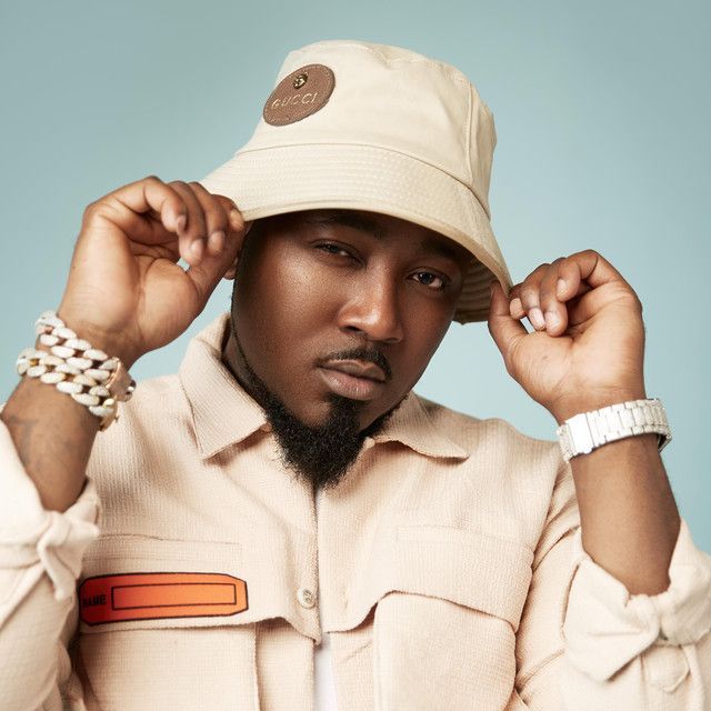 Man in cream outfit adjusting beige bucket hat against a pale blue background