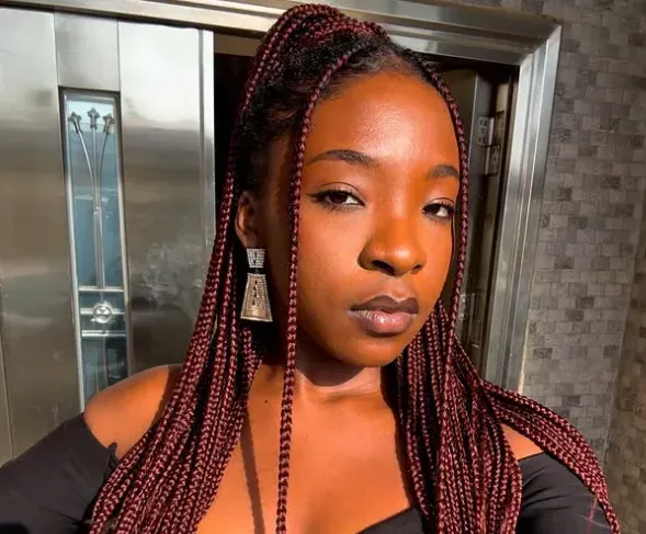 Woman with long braided hair posing indoors near an elevator, wearing a black top and square earrings.