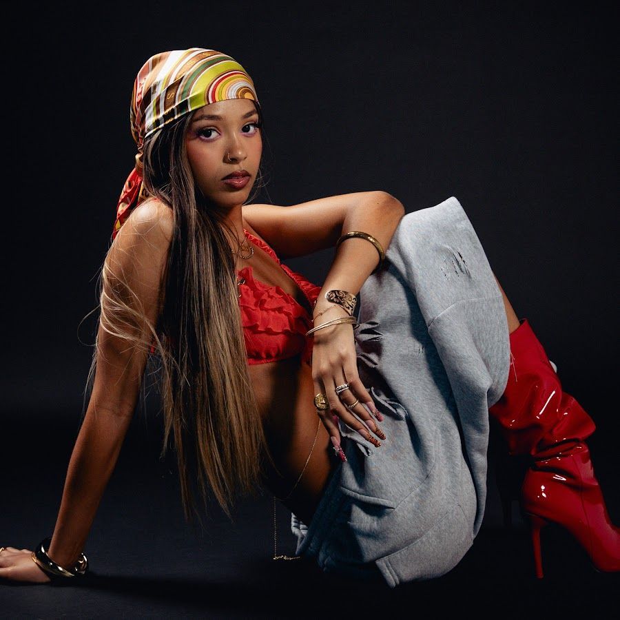 Person in a red top, gray pants, and patterned headscarf posing seated against a dark background.