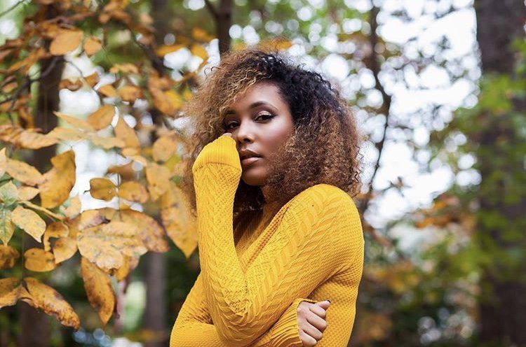 Woman in a yellow sweater standing among autumn trees, posing thoughtfully.