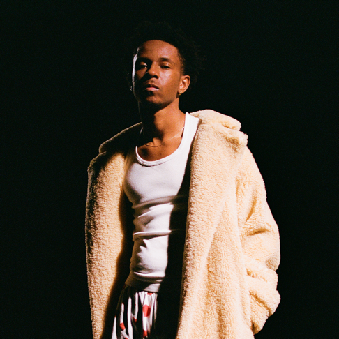 Person in a beige jacket over a white tank top, standing against a dark background with hands in pockets