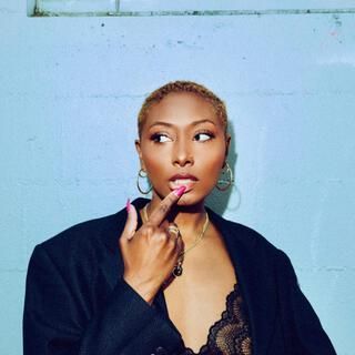 Person in black blazer and lace top, touching lips thoughtfully against a light blue wall.