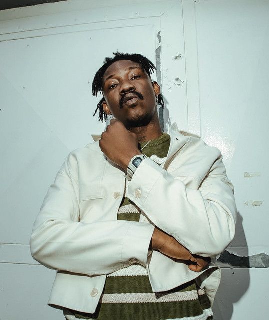 Person in a white jacket and striped sweater poses with arms crossed against a light wall.