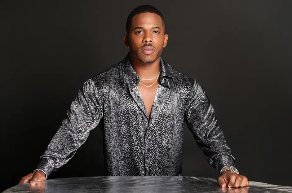 Man in a silver patterned shirt stands behind a table against a dark background.