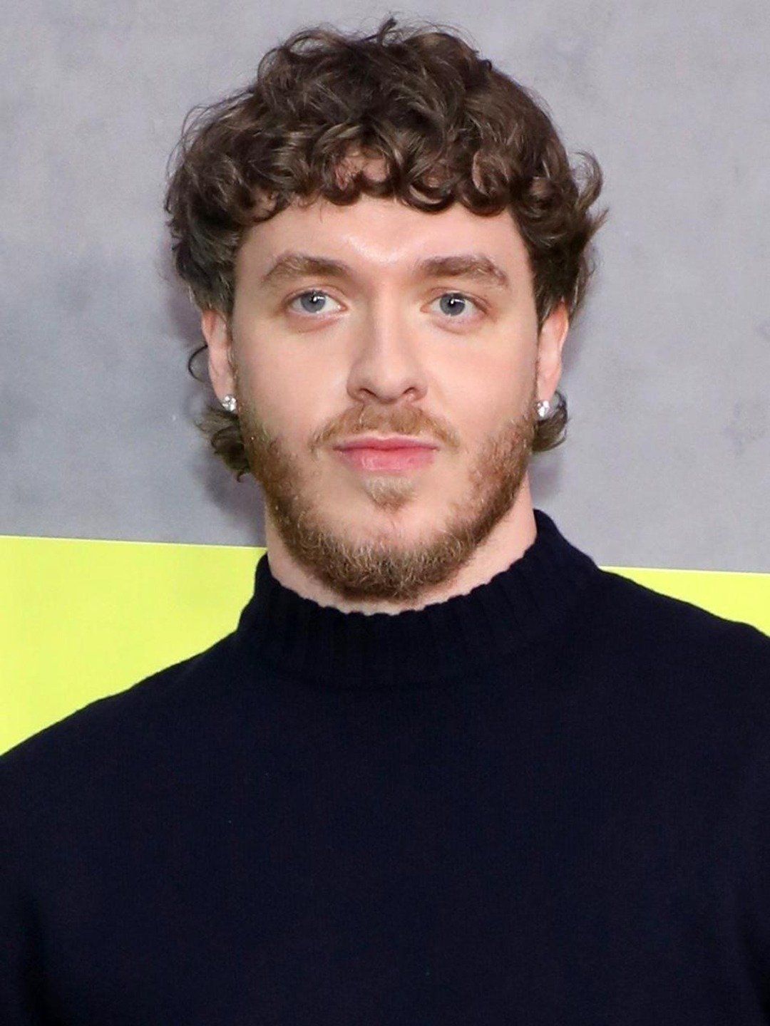 Portrait of a person with curly hair and beard, wearing a black sweater against a yellow background