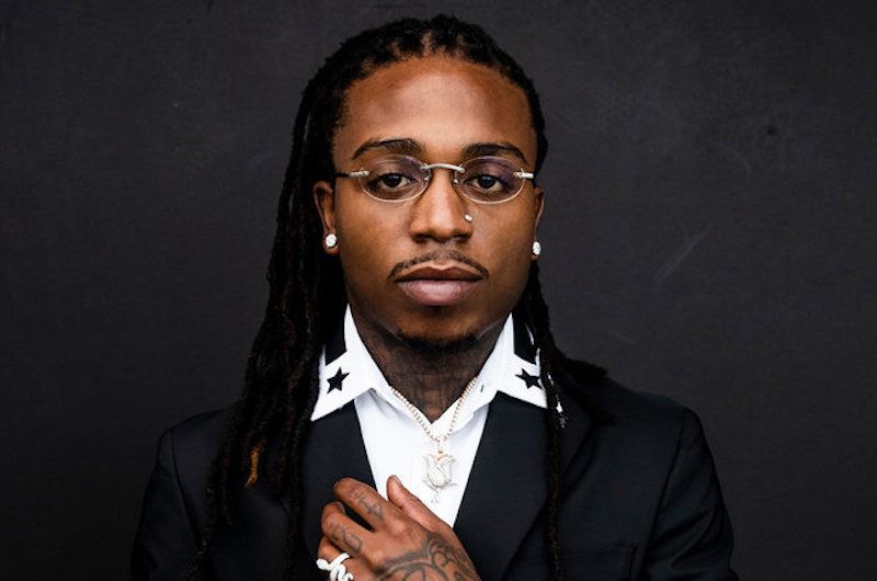 Portrait of a person in a black suit and white shirt, wearing glasses against a dark background