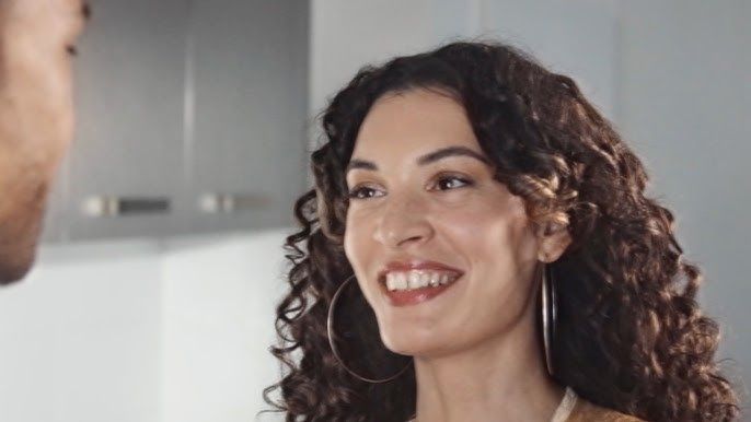 Smiling woman with curly dark hair in a kitchen, facing a person at the left edge