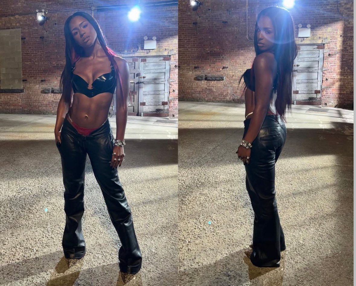Woman posing in a black outfit outdoors at night, with brick walls and bright lights behind her.