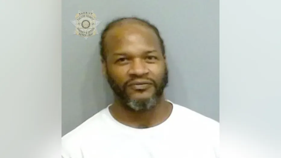 Man standing against a gray wall with a visible police emblem, wearing a white shirt and looking at the camera.