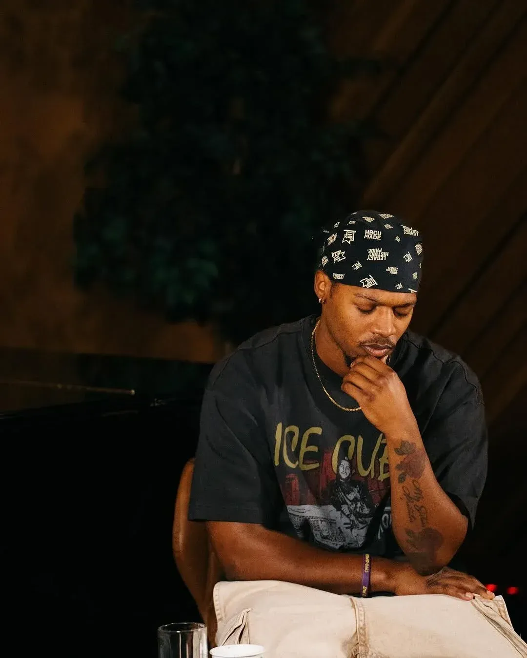 A man in a black shirt and patterned bandana sits thoughtfully on a chair in a dim studio.