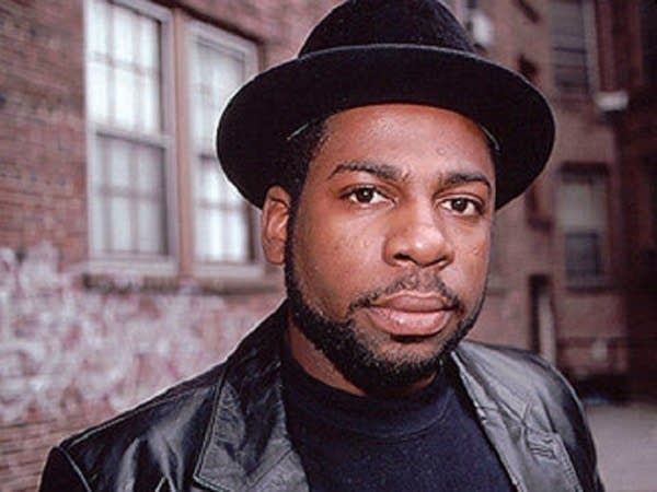 Man wearing a black hat and leather jacket standing outdoors in front of brick buildings