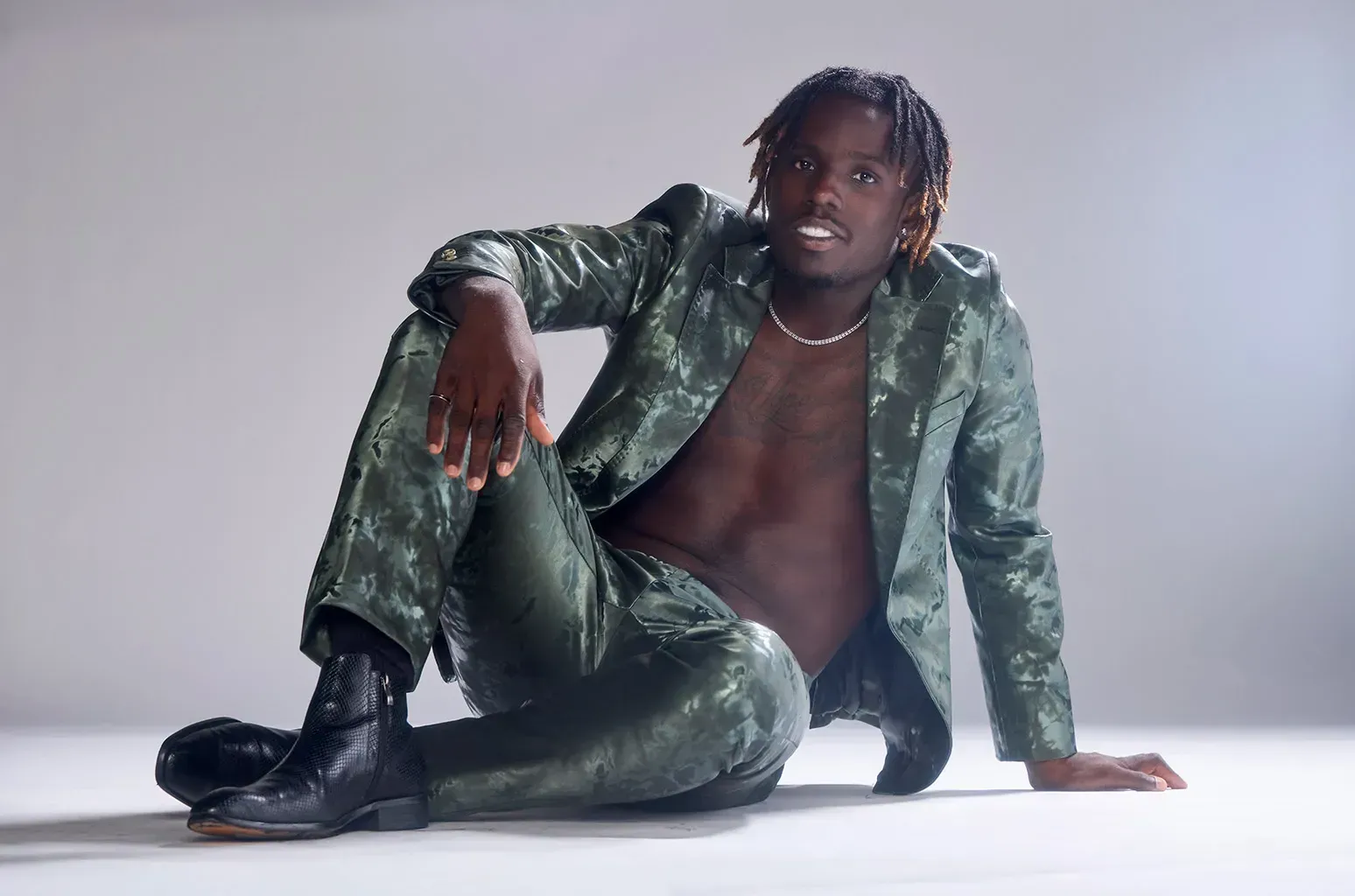 Person in a shiny green outfit posing seated on a white studio floor against a plain background