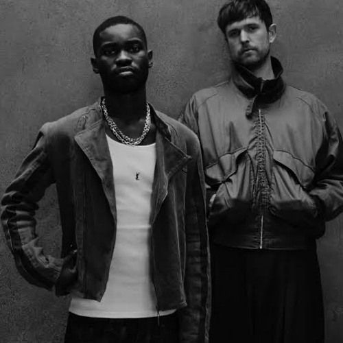 Two men posing in a studio, one in a dark jacket and white shirt, the other in a gray jacket.