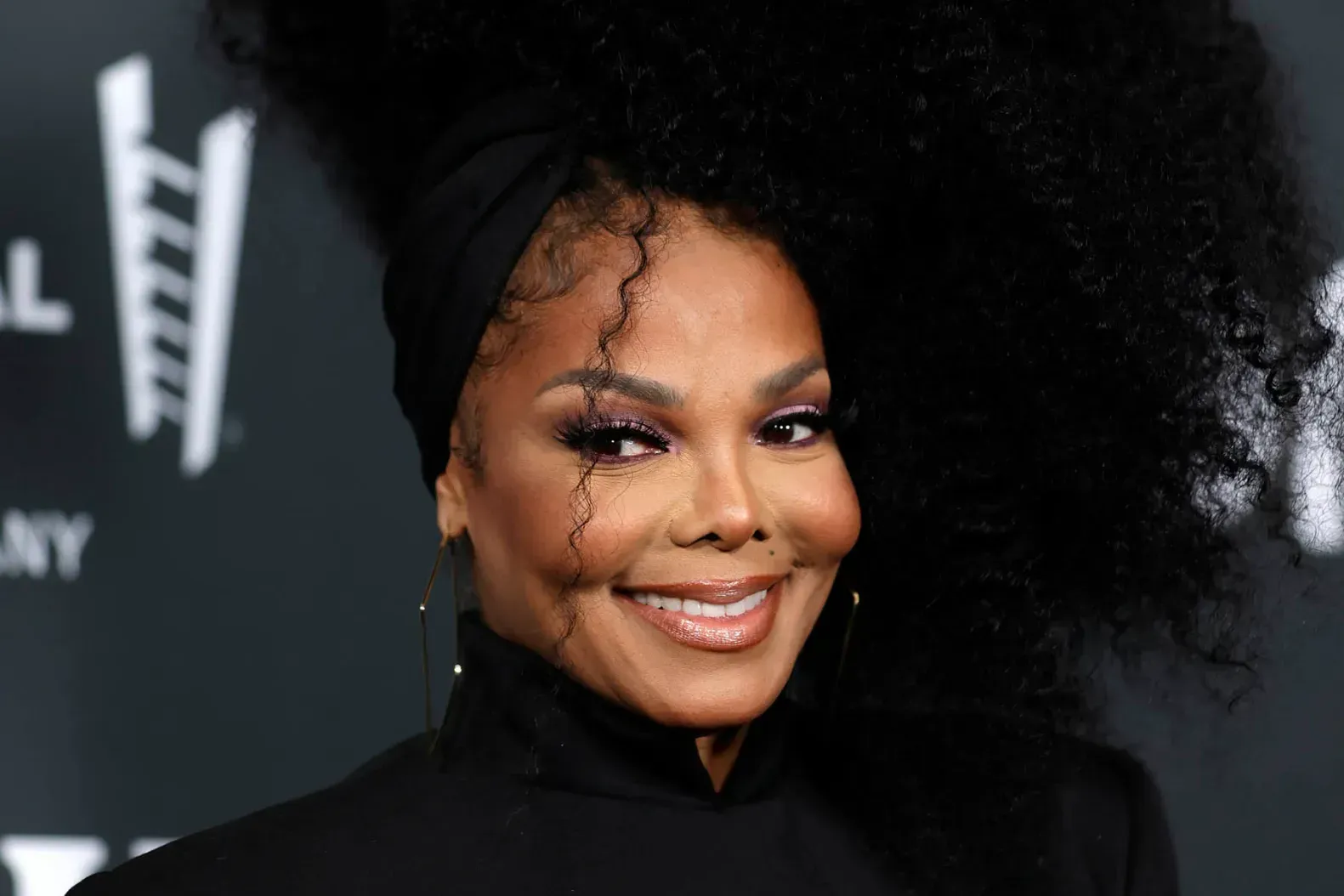 Smiling woman in black outfit at a red carpet event with a dark backdrop
