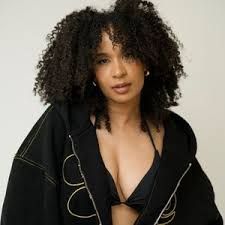 Woman with curly hair wearing a black jacket and top, posing against a plain light background