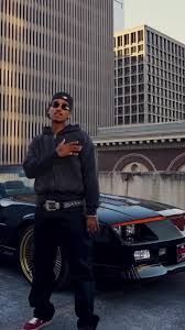 Person in black outfit standing by a black car with city skyscrapers in the background.
