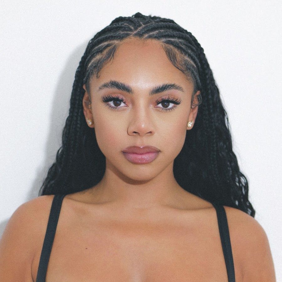 Portrait of a woman with braided dark hair, wearing a black strappy top, against a light background