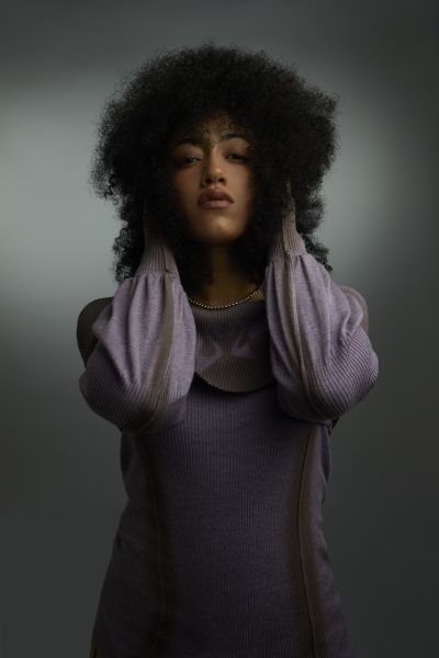 Portrait of a person in a lavender sweater, hands near head, against a dark gray background.