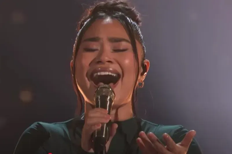 Singer passionately performing into a microphone on a dark stage, eyes closed and hand raised.