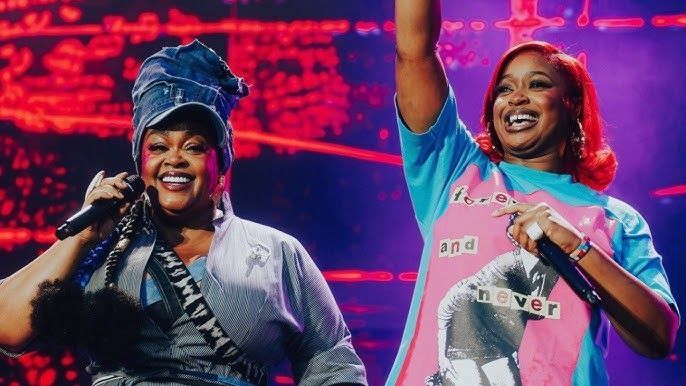 Two smiling performers onstage under pink lights, one waving with a microphone, the other in a blue headwrap and gray top