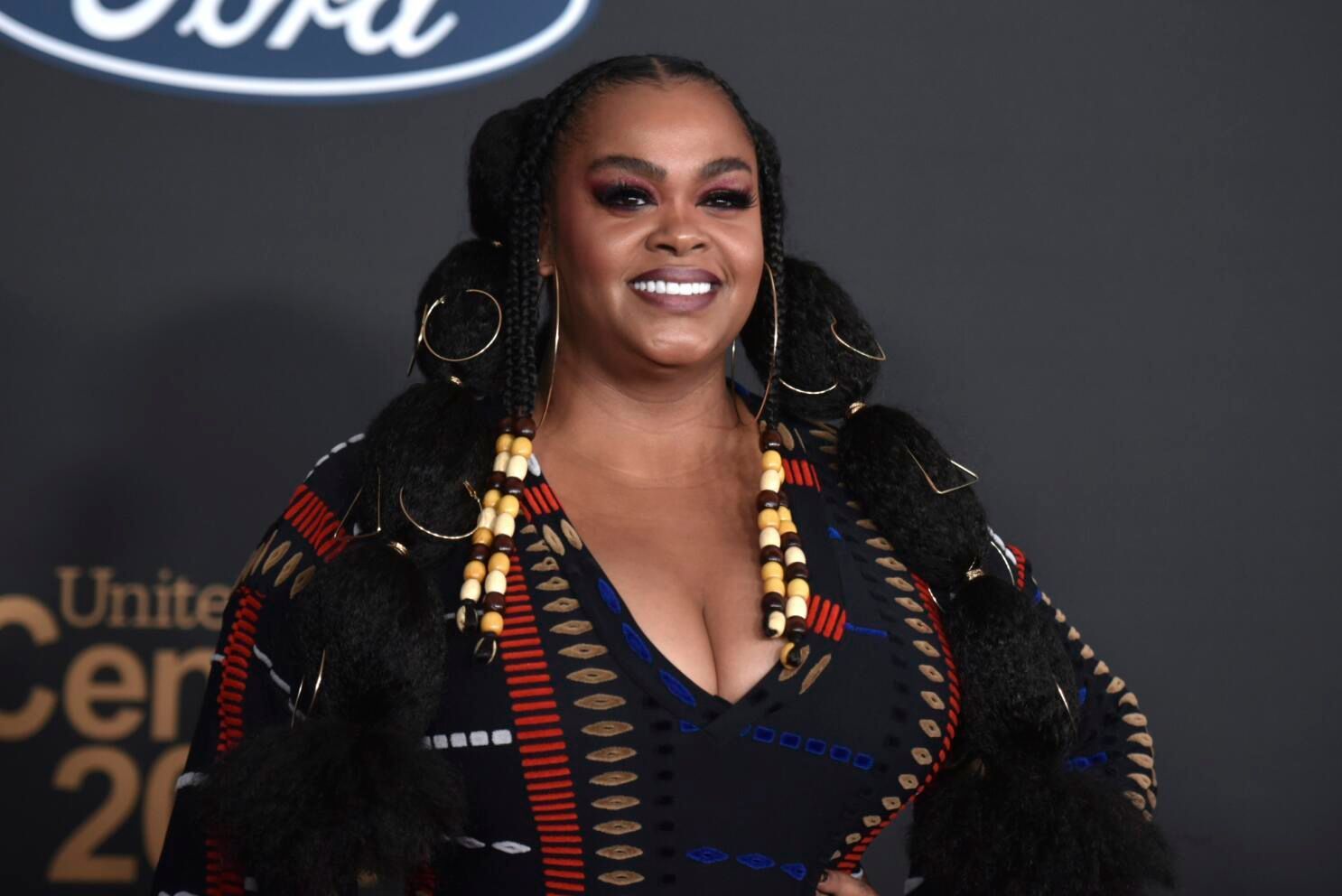 Woman smiling on a red carpet in a black dress with colorful trim and large hoop earrings