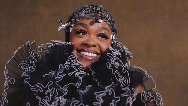 Smiling woman in a black ruffled outfit and jeweled headpiece against a brown background
