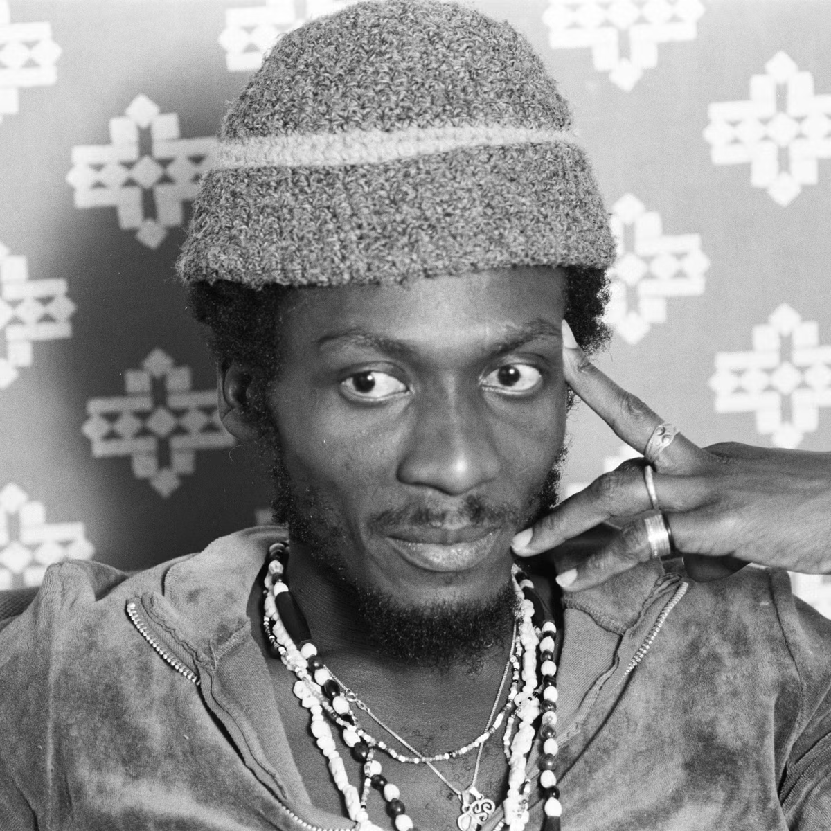 Black-and-white portrait of a person in a knit hat, jewelry, and a denim jacket, resting a hand on their face