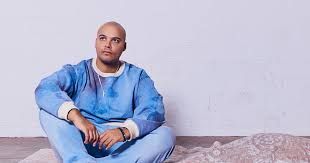 Person in a light blue outfit sitting on the floor against a wall, looking off to the side.