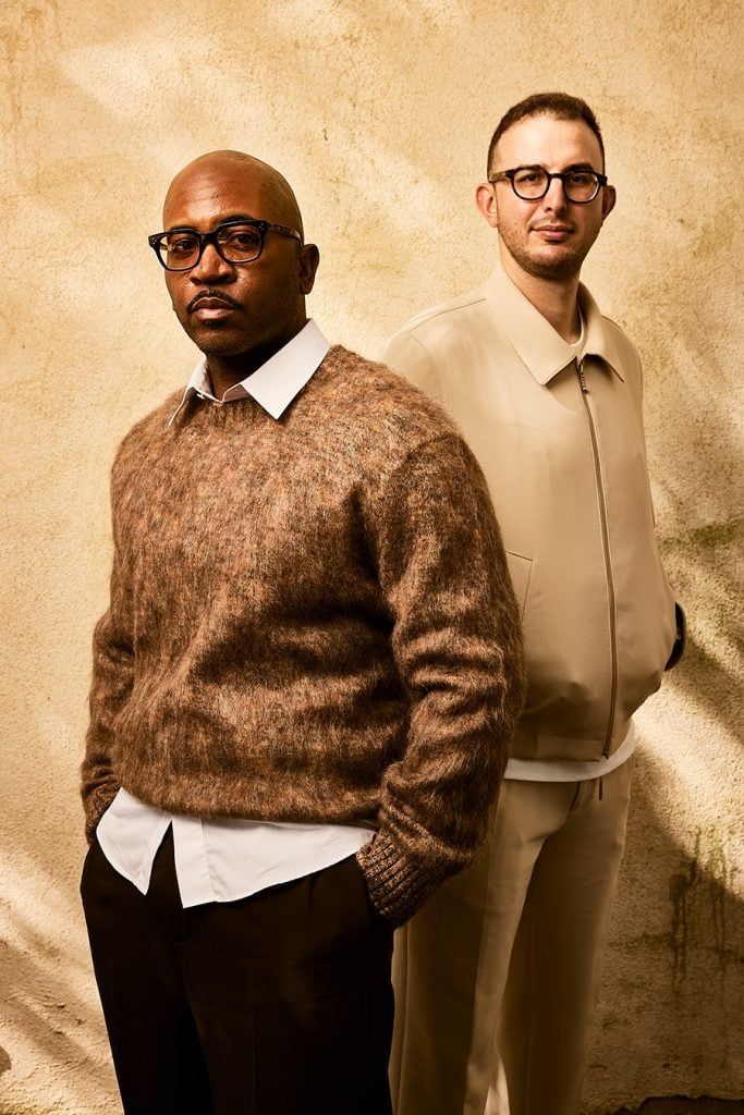 Two men standing back to back against a textured beige wall, one in a brown sweater and one in a cream sweater.