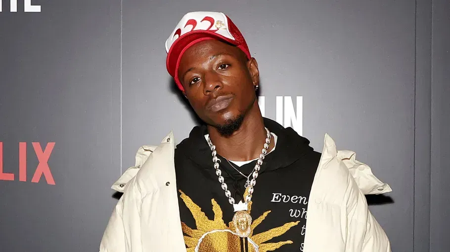Man posing at a Netflix event, wearing a red cap, black shirt, and light jacket against a gray backdrop