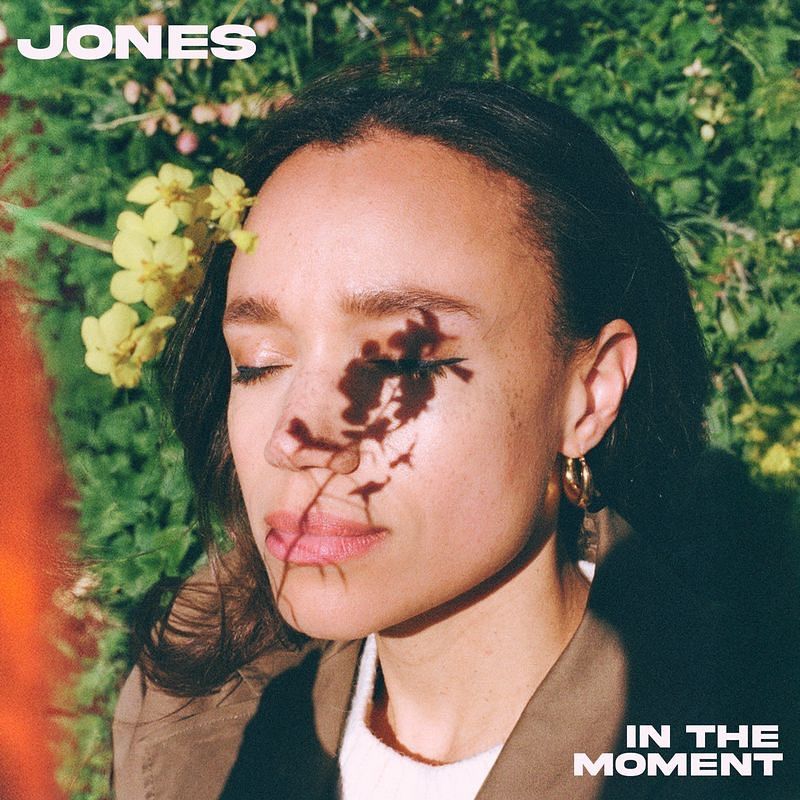 Woman with eyes closed among flowers and foliage, with “JONES” and “IN THE MOMENT” text.