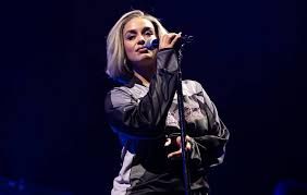 Singer at a microphone on a dark stage, wearing a black and gray outfit with blonde hair.