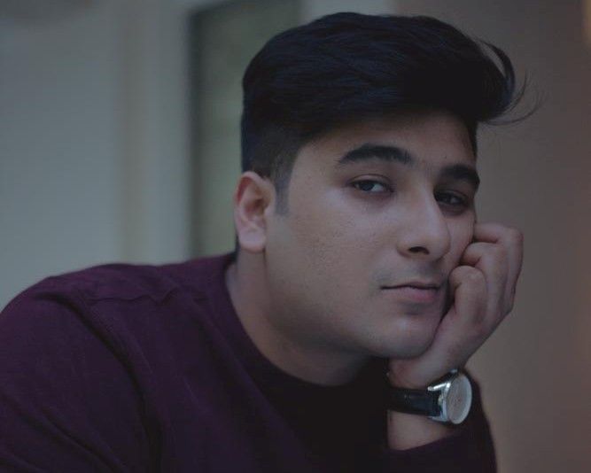 Person with dark hair and maroon shirt rests their head on their hand in a dim, neutral setting.