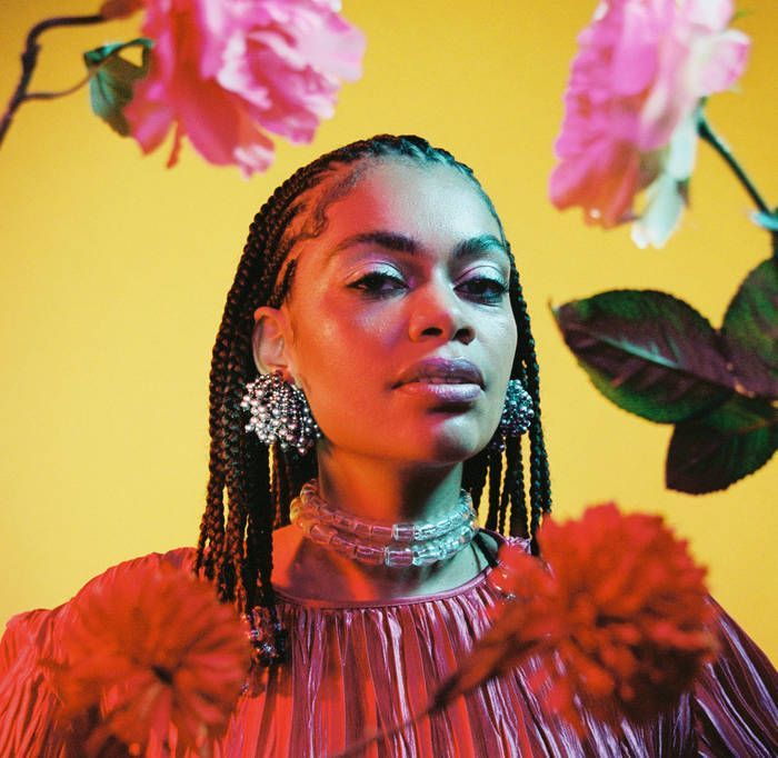 Portrait of a person with braided hair and floral earrings against a bright yellow background with pink flowers