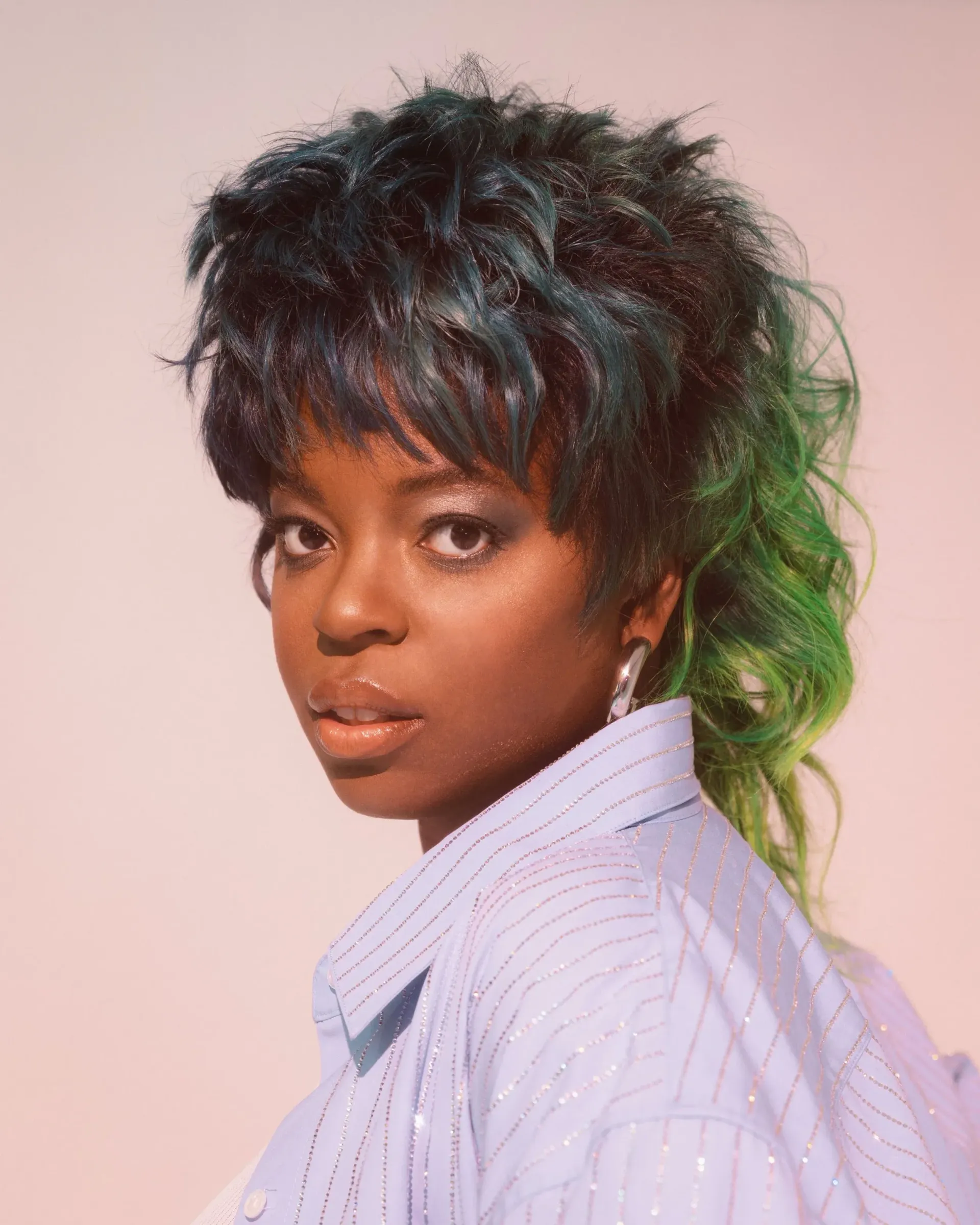 Portrait of a person with short dark hair and green highlights, wearing a light striped shirt, against a pink background