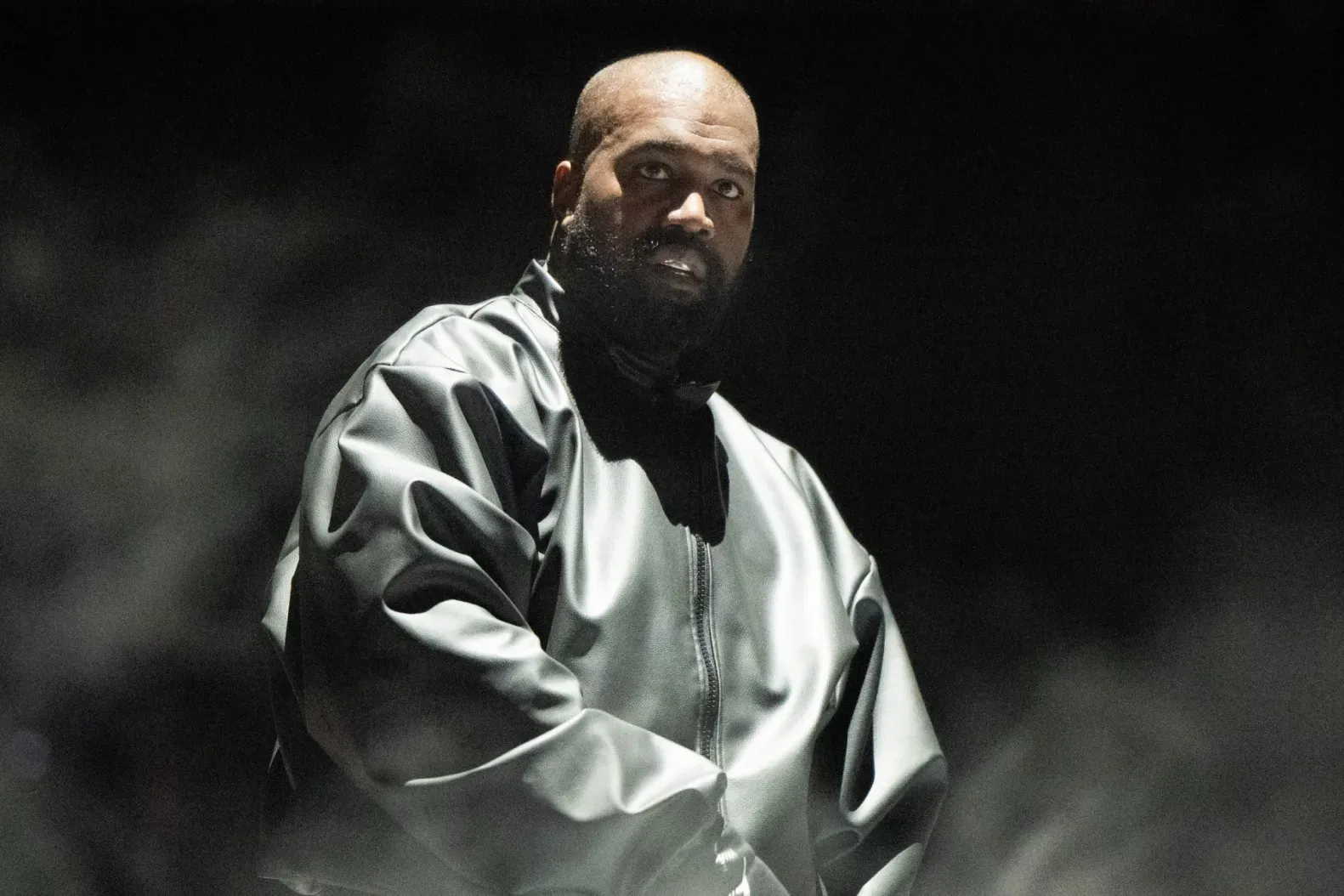 Man in a shiny jacket stands in a dark, smoky setting, looking directly ahead.