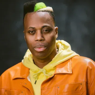 Person in an orange jacket and yellow hoodie, with a short green-and-black hairstyle, against a dark background.