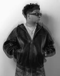 Person in sunglasses and a black leather jacket standing against a plain wall, looking to the side.