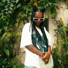 Person with long braids and white sunglasses standing in greenery, wearing a white shirt and looking down