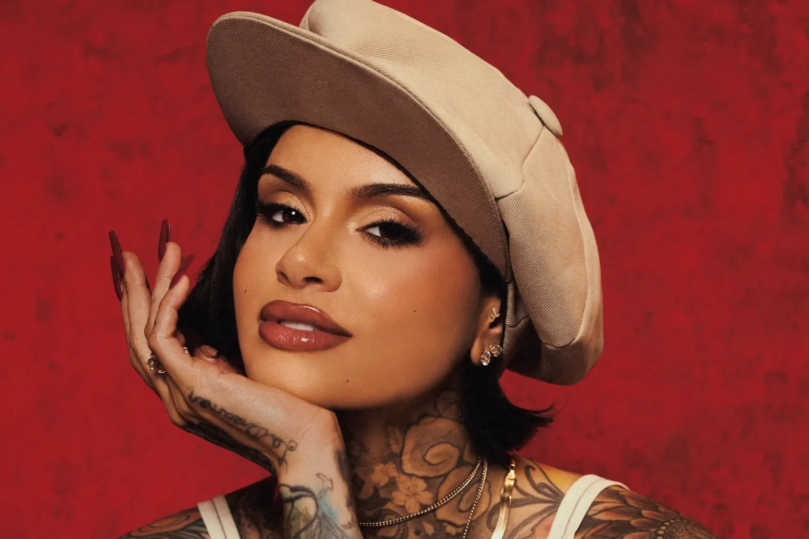 Portrait of a person in a beige hat against a red background, resting a hand on their cheek