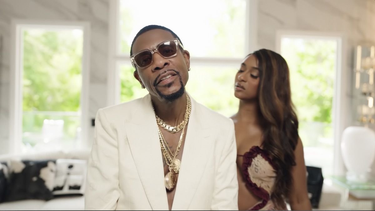 A man in a white suit and sunglasses stands before a woman in a maroon dress in a bright room.
