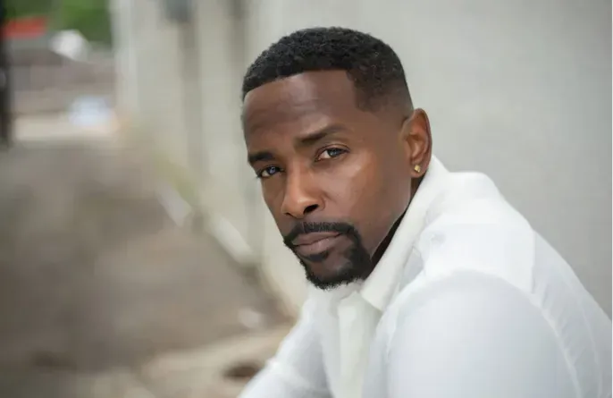 Man in a white shirt leaning outdoors, looking at the camera with a calm expression.