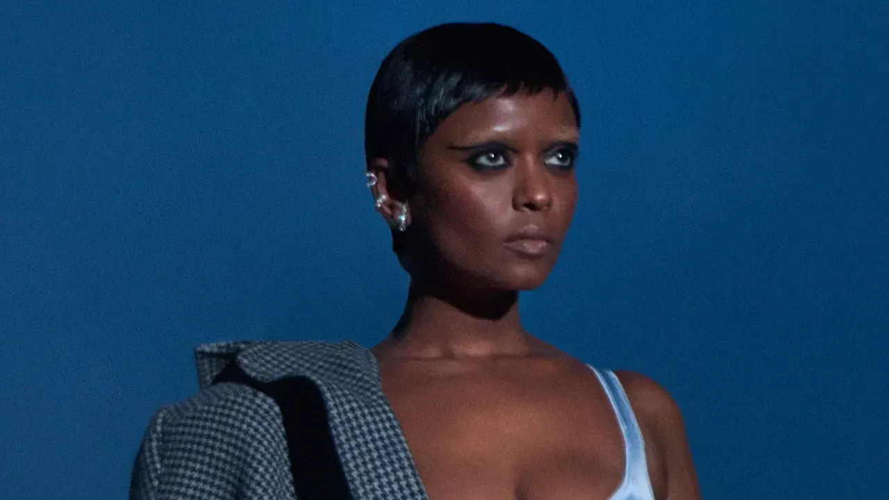 Portrait of a person with short hair against a blue background, wearing a light blue top and checkered jacket.