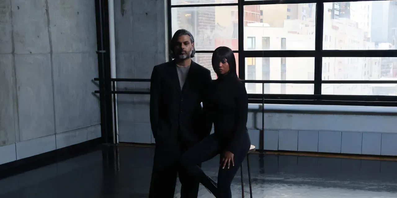 Two people in dark clothing standing in a modern room with large windows.